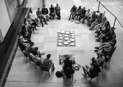 A black and white photo taken from above of a drum circle about to begin. Students are sitting in a circle listening to the instructor.