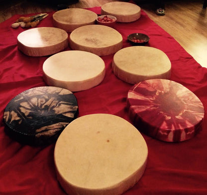 Nine hand drums are laid out on the floor on red fabric. Two have intricate patterns, seven are solid tan coloured. 